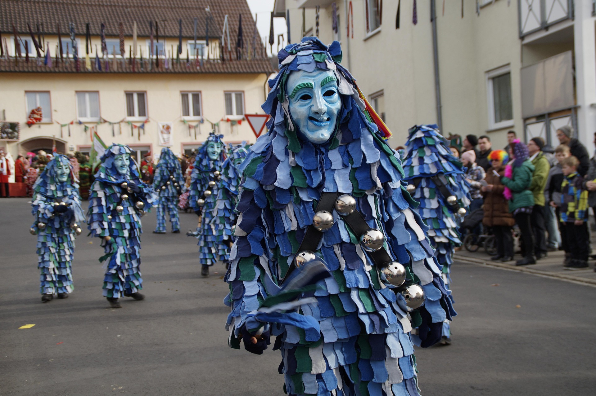 Gruppe von Narren in blauem Häs mit Stoffblättern und Schellen beim Umzug, Zuschauer am Straßenrand